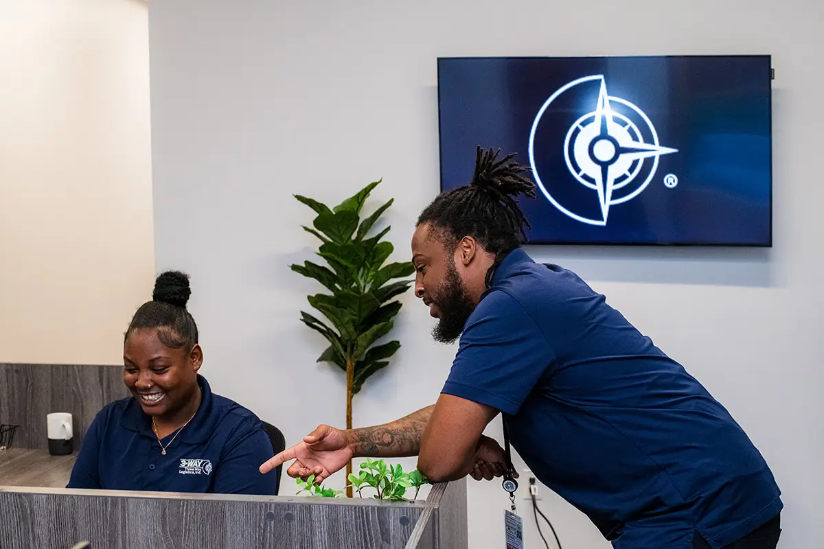 Three Way Logistics team members collaborating at a reception desk, with one employee pointing at a screen while another smiles, and the company logo displayed on a monitor in the background