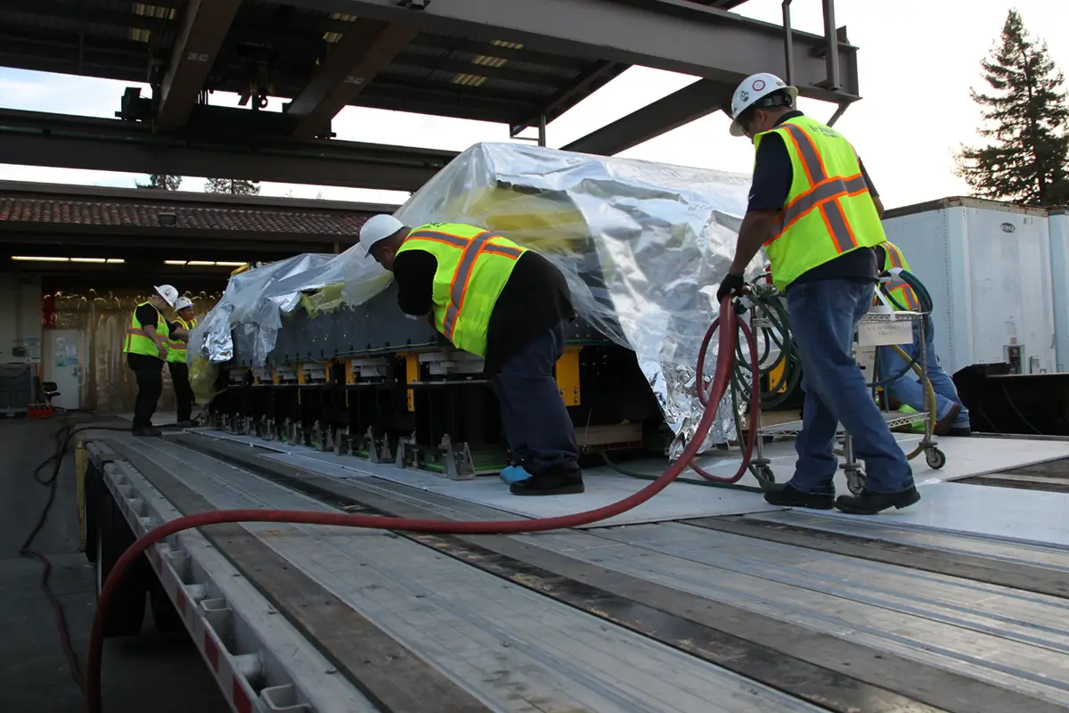 Workers in safety vests and hard hats carefully unloading and handling large industrial equipment wrapped in protective plastic on a flatbed trailer